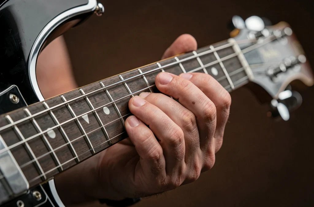 Gros plan sur les doigts d'un guitariste formant l'accord B7 sur le manche d'une guitare électrique noire et chrome